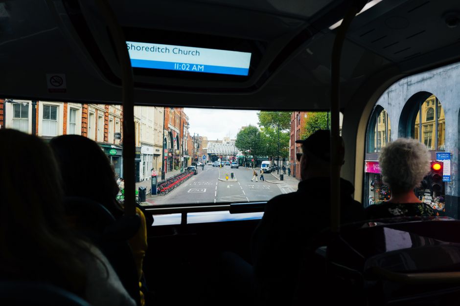 View from inside a bus during an early morning home relocation journey on the route from Old Street to Hoxton, with three passengers seated and observing an urban street scene through the front windsh