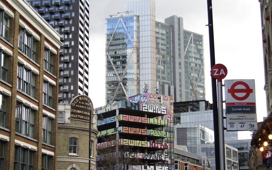 A light blue transport van is positioned on the street in front of an ornate, multi-story brick building in Shoreditch, London. The van appears to be engaged in the loading process, with its side door